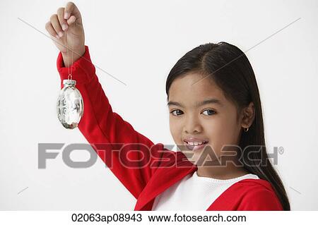 Stock Image - Girl holding up Christmas ornament looking at camera. Fotosearch