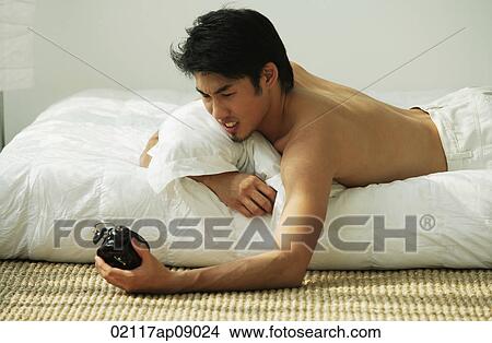 Picture - Young man lying in bed, holding alarm clock. Fotosearch
