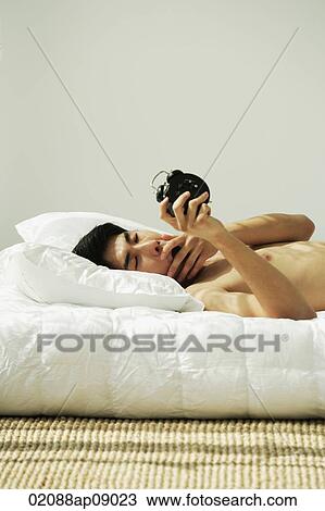 Young man lying in bed, yawning and holding alarm clock View Large Photo Image Stock Image - Young man lying in bed, yawning and holding alarm clock. Fotosearch