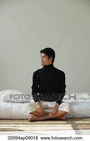 Stock Photo - Young man sitting on bed, looking sideways. Fotosearch