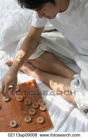 Young man sitting on bed, playing chinese chess View Large Photo Image Stock Photo - Young man sitting on bed, playing chinese chess. Fotosearch