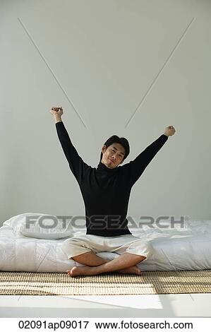 Stock Photo - Young man sitting on bed, stretching arms. Fotosearch