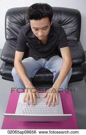 Stock Photograph - Young man sitting on couch using laptop. Fotosearch