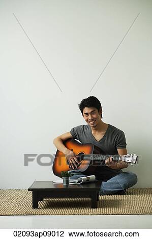 Stock Image - Young man sitting on floor playing guitar smiling at camera. Fotosearch