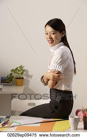 Stock Photography - Chinese fashion designer sitting on desk, smiling at camera. Fotosearch