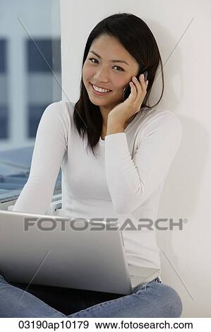 Young Asian woman talking on phone while looking at laptop View Large Photo Image Stock Photo - Young Asian woman talking on phone while looking at laptop. Fotosearch