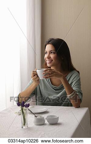 Stock Photograph - young woman holding coffee cup and looking out window smiling. Fotosearch