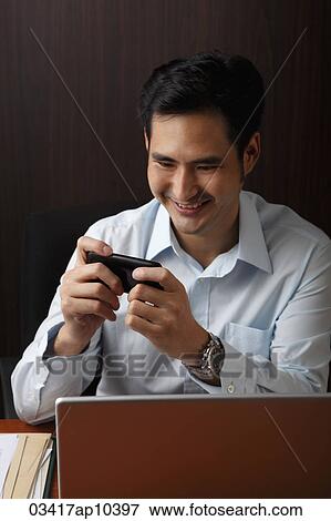 man sitting at desk and looking at phone and smiling View Large Photo Image Stock Photo - man sitting at desk and looking at phone and smiling. Fotosearch