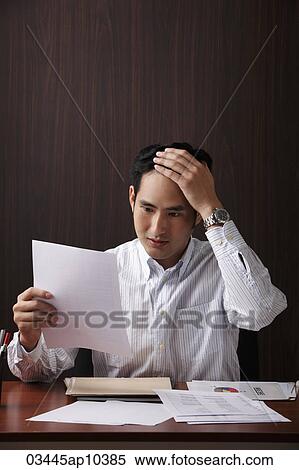Stock Photography - man sitting at desk holding paper looking stressed. Fotosearch