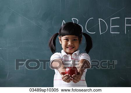 Stock Photography - young girl holding out apple in front of chalkboard. Fotosearch