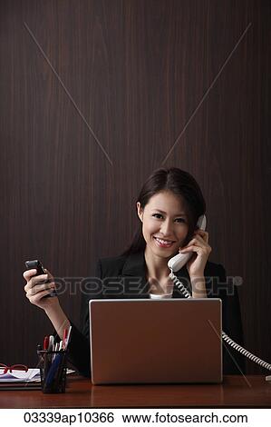 Stock Photograph - Young woman sitting at her desk holding her phones. Fotosearch