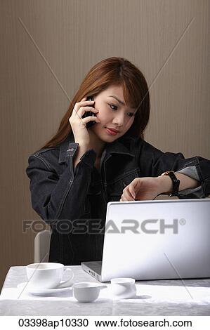 Young woman sitting at table with lap top, talking on phone, looking at watch View Large Photo Image Stock Image - Young woman sitting at table with lap top, talking on phone, looking at watch. Fotosearch