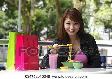 Stock Photography - Young woman sitting outside eating noodles. Fotosearch