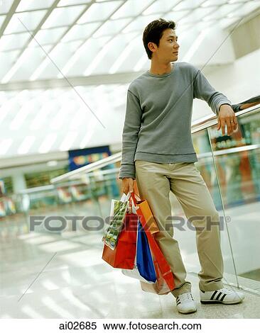 Stock Photography - Casual dressed man holding shopping bags in mall leaning on railing.. Fotosearch