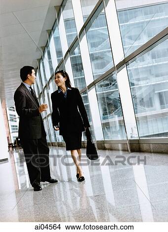 Picture - Executive pair walking down hallway talking, large windows in background.. Fotosearch