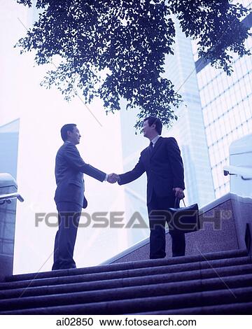 Stock Image - Executives shaking hands at top of stairs, buildings in background.. Fotosearch