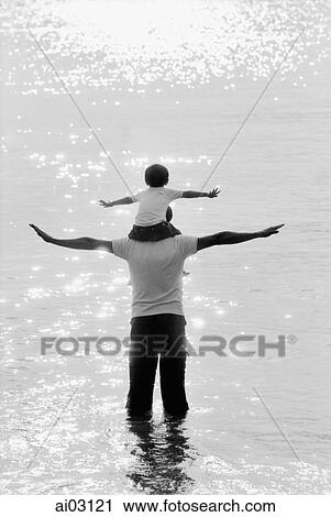 Stock Image - Father and son wading in water, son on father's shoulders, arms stretched. Fotosearch