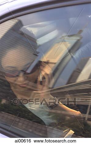 Stock Photograph - Female executive with sunglasses using cellular phone in car, skyline reflecting in window. Fotosearch