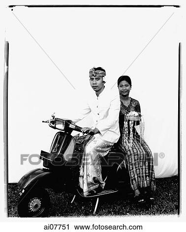 Stock Image - Indonesia, Bali, Ubud, Balinese wedding couple in ceremonial dress, sitting on motor scooter, woman carrying offering.. Fotosearch