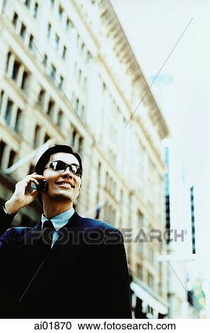 Stock Image - Male executive wearing sunglasses using cellular phone, building in background ( high grain). Fotosearch