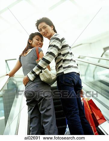 Stock Image - Man and woman carrying shopping bags going upward on escalator looking back.. Fotosearch