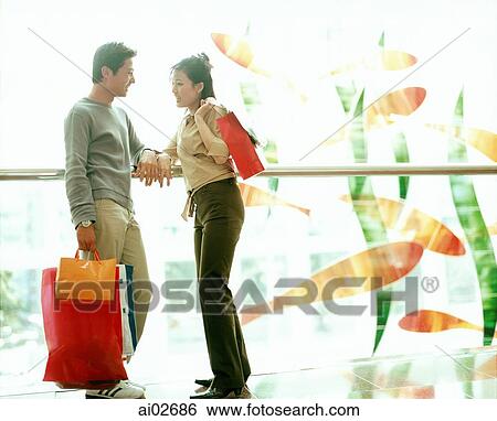 Stock Photograph - Man and woman holding shopping bags talking and leaning on railing.. Fotosearch