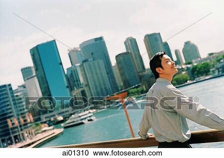 Stock Image - Man leaning on railing overlooking bay, skyline in background. Fotosearch