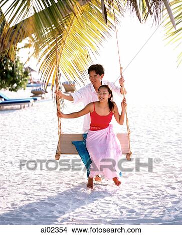 Man pushing woman on swing under coconut tree looking off camera, sea in background View Large Photo Image Picture - Man pushing woman on swing under coconut tree looking off camera, sea in background. Fotosearch
