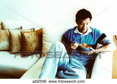Man sitting on sofa eating View Large Photo Image Stock Photograph - Man sitting on sofa eating. Fotosearch