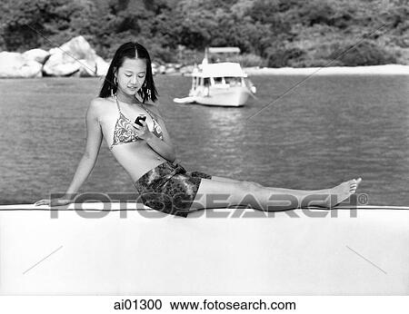 Stock Image - Teenage girl in bikini using cellular phone, boat in background. Fotosearch