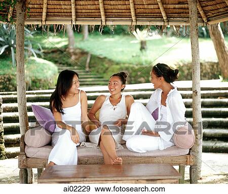 Stock Photo - Three women wearing white, sitting together on bench talking. Fotosearch