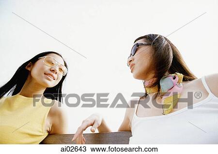 Two young women with sunglasses sitting and talking, low angle view View Large Photo Image Stock Image - Two young women with sunglasses sitting and talking, low angle view. Fotosearch