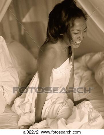 Stock Image - Woman draped in sheet sitting in bed, laughing. Fotosearch