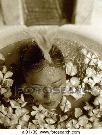 Stock Image - Woman in bath with floating flowers, head under running water. Fotosearch