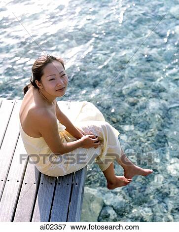 Stock Photo - Woman in yellow dress sitting on dock, water in background, portrait, elevated position. Fotosearch