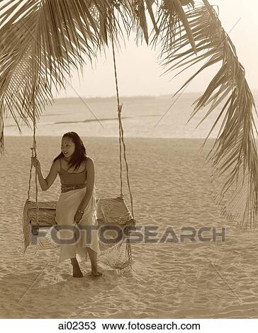 Stock Image - Woman sitting on swing under coconut tree looking off camera, sea in background. Fotosearch