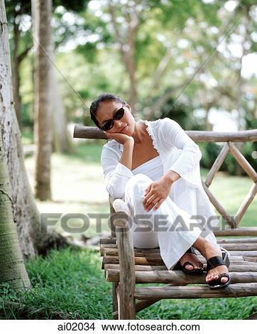 Picture - Woman sitting on wooden bench, trees behind. Fotosearch