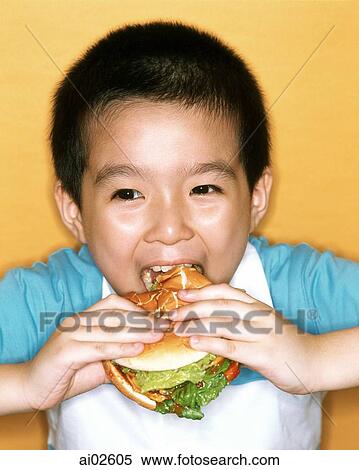 Stock Photography - Young boy eating large hamburger, yellow background.. Fotosearch