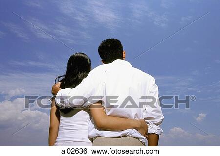 Young couple embracing with blue sky in background, rear view View Large Photo Image Stock Photograph - Young couple embracing with blue sky in background, rear view. Fotosearch