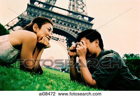 Stock Image - Young couple lying on grass, young woman posing for camera, smiling, Eiffel Tower in background. (high-grained). Fotosearch