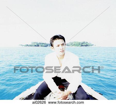 Stock Photograph - Young man sitting on boat, island in background, portrait. Fotosearch