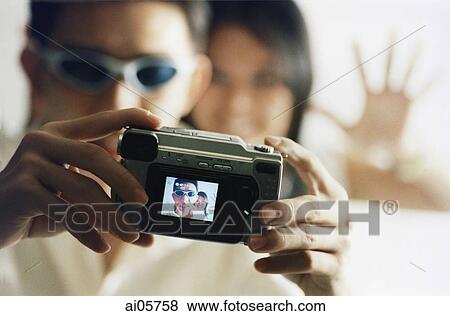 Young man using digital camera with woman waving in background ( high grain) View Large Photo Image Stock Photo - Young man using digital camera with woman waving in background ( high grain). Fotosearch