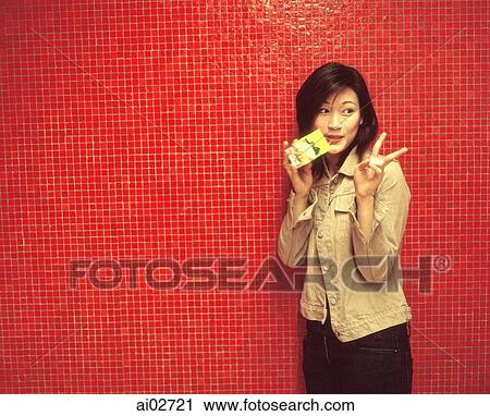 Stock Image - Young woman making a peace sign, drinking a packet drink, red background.. Fotosearch