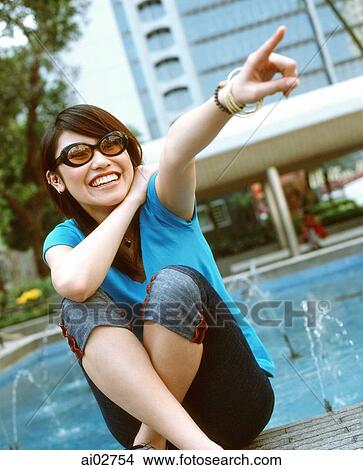 Young woman wearing sunglasses sitting and pointing. View Large Photo Image Picture - Young woman wearing sunglasses sitting and pointing.. Fotosearch