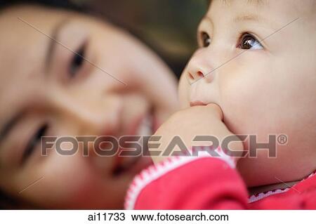 Stock Image - Baby girl with hand in mouth, mother looking at her. Fotosearch