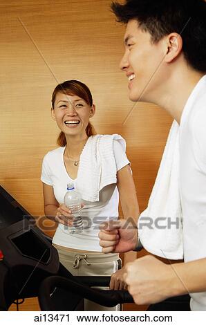 Stock Image - Couple in gym, man on treadmill, woman holding bottle of water. Fotosearch