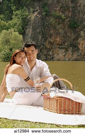 Couple sitting by a lake, with picnic basket, looking at camera View Large Photo Image Picture - Couple sitting by a lake, with picnic basket, looking at camera. Fotosearch
