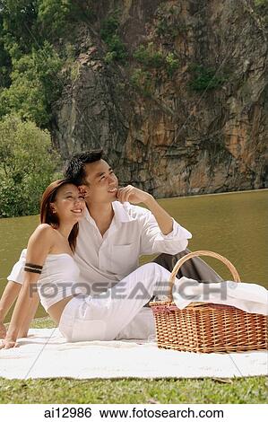 Couple sitting by a lake, with picnic basket, looking away View Large Photo Image Stock Photograph - Couple sitting by a lake, with picnic basket, looking away. Fotosearch