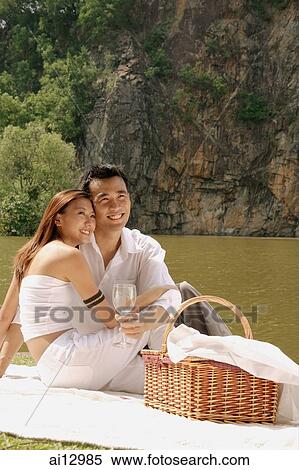 Stock Photography - Couple sitting by a lake, with picnic basket, looking away. Fotosearch