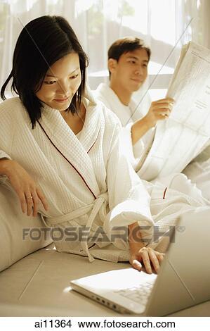 Couple sitting down, man reading newspaper, woman using laptop View Large Photo Image Picture - Couple sitting down, man reading newspaper, woman using laptop. Fotosearch